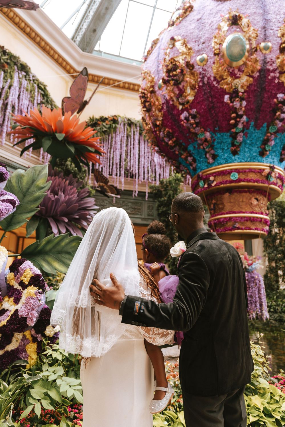 Bride and groom stand among vibrant floral displays inside Bellagio Conservatory, with a colorful hot air balloon backdrop.