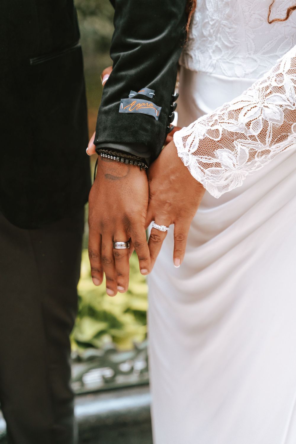 Close-up of newlyweds holding hands showing wedding rings, bride in lace dress groom in black suit.