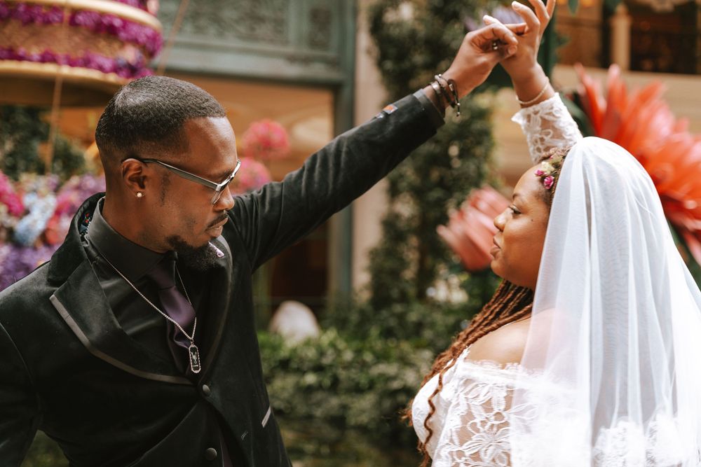 A groom in a black suit twirls his bride in a white lace wedding dress and veil outdoors.