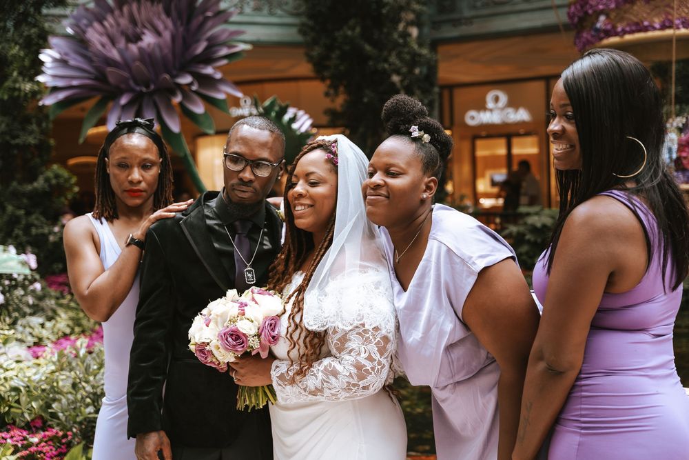 Bride in white gown holds bouquet surrounded by smiling family members in purple dresses at floral wedding venue.