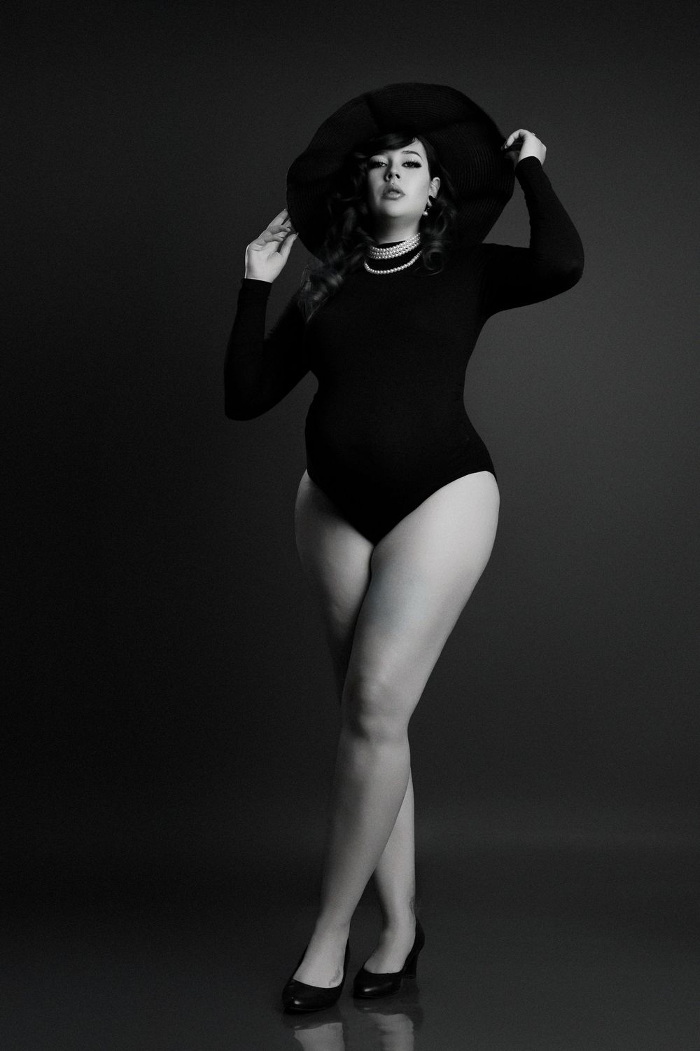 Black and white photo of a woman in a bodysuit and wide-brim hat posing confidently in heels.