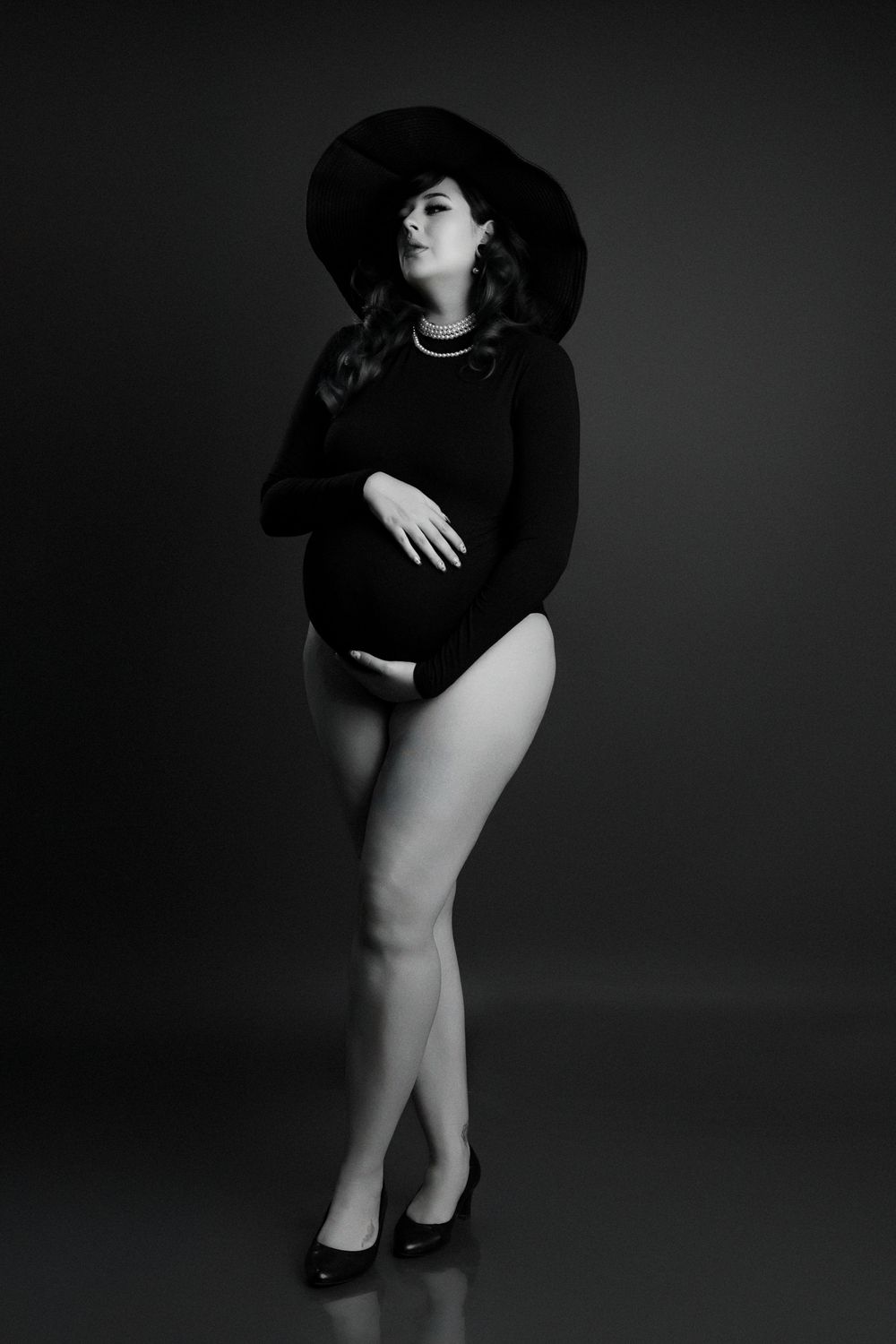Black and white maternity photo of pregnant woman in black bodysuit and wide-brim hat posing elegantly in studio.