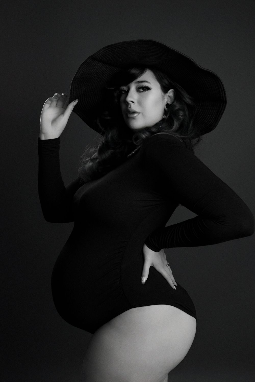 Black and white photo of a woman in a bodysuit and wide-brim hat posing elegantly in a studio setting.
