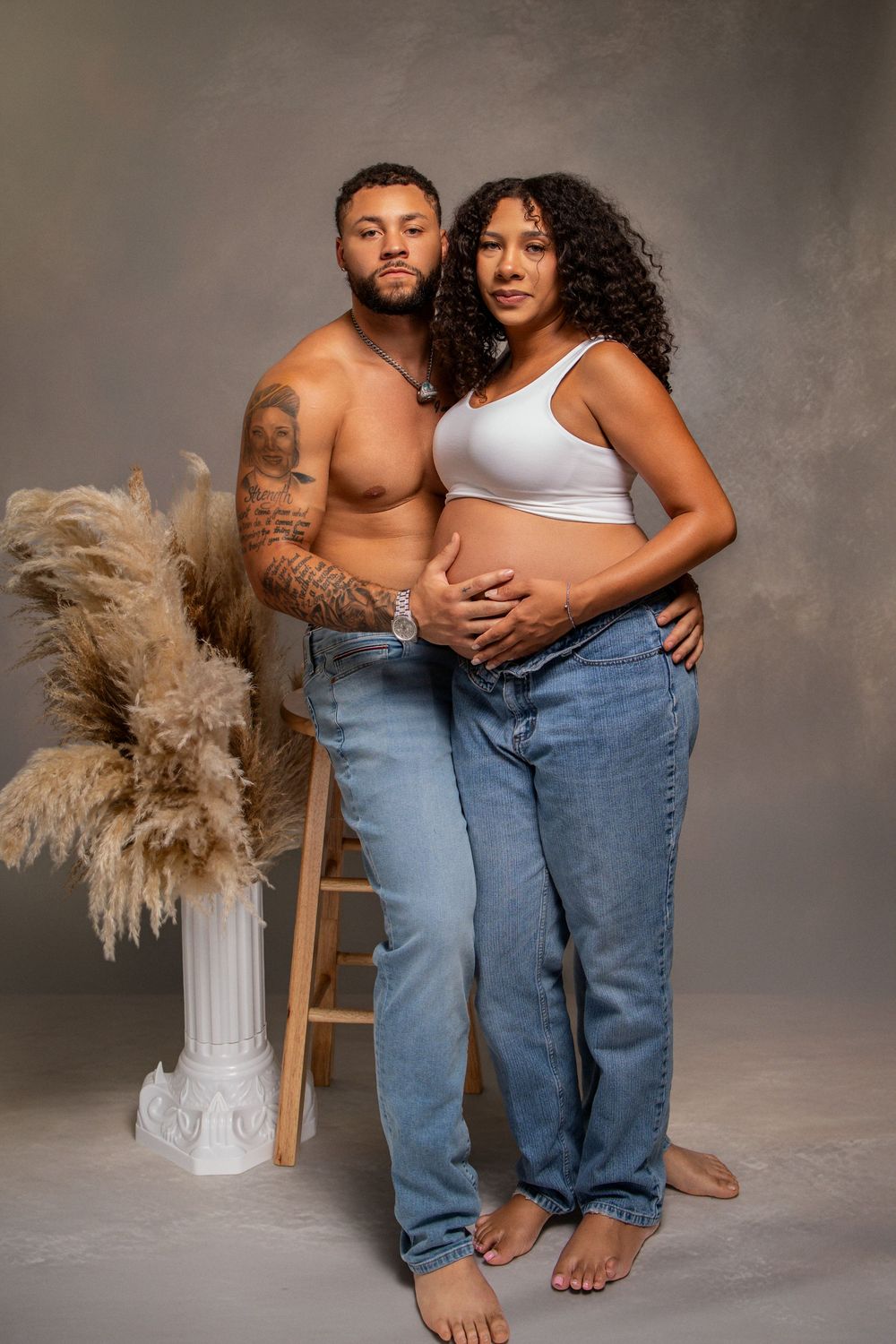 A shirtless tattooed man embraces a pregnant woman in a white crop top and jeans in a studio maternity photo shoot.
