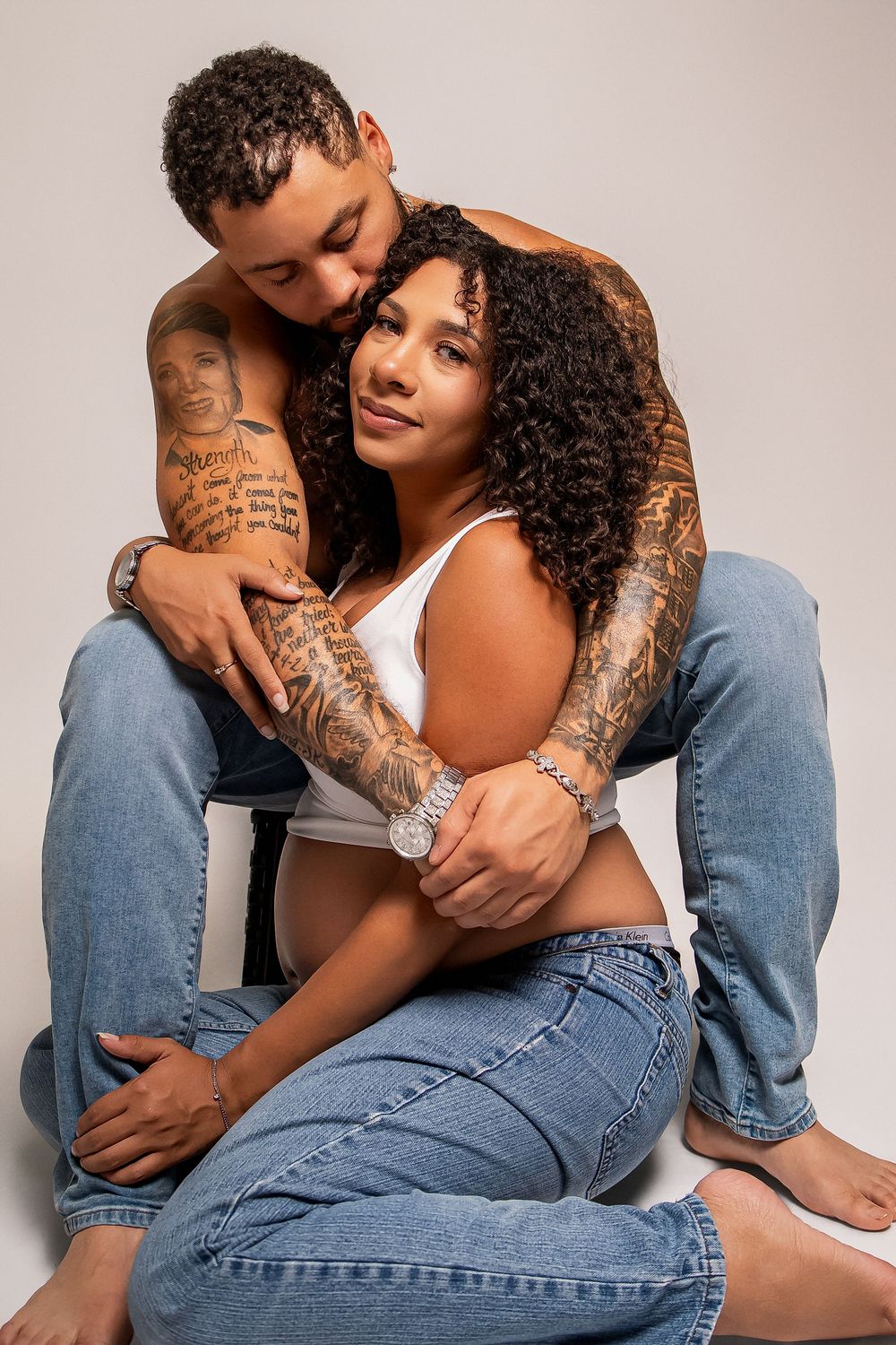 Tattooed couple in jeans embracing in studio, woman in white crop top smiling at camera while man kisses her cheek.