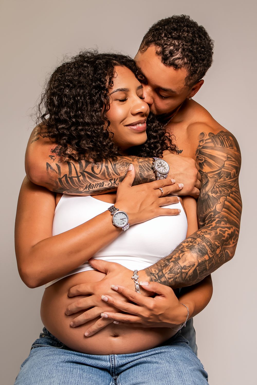 Tattooed couple embracing tenderly, pregnant woman in white crop top and jeans, man kissing her head.