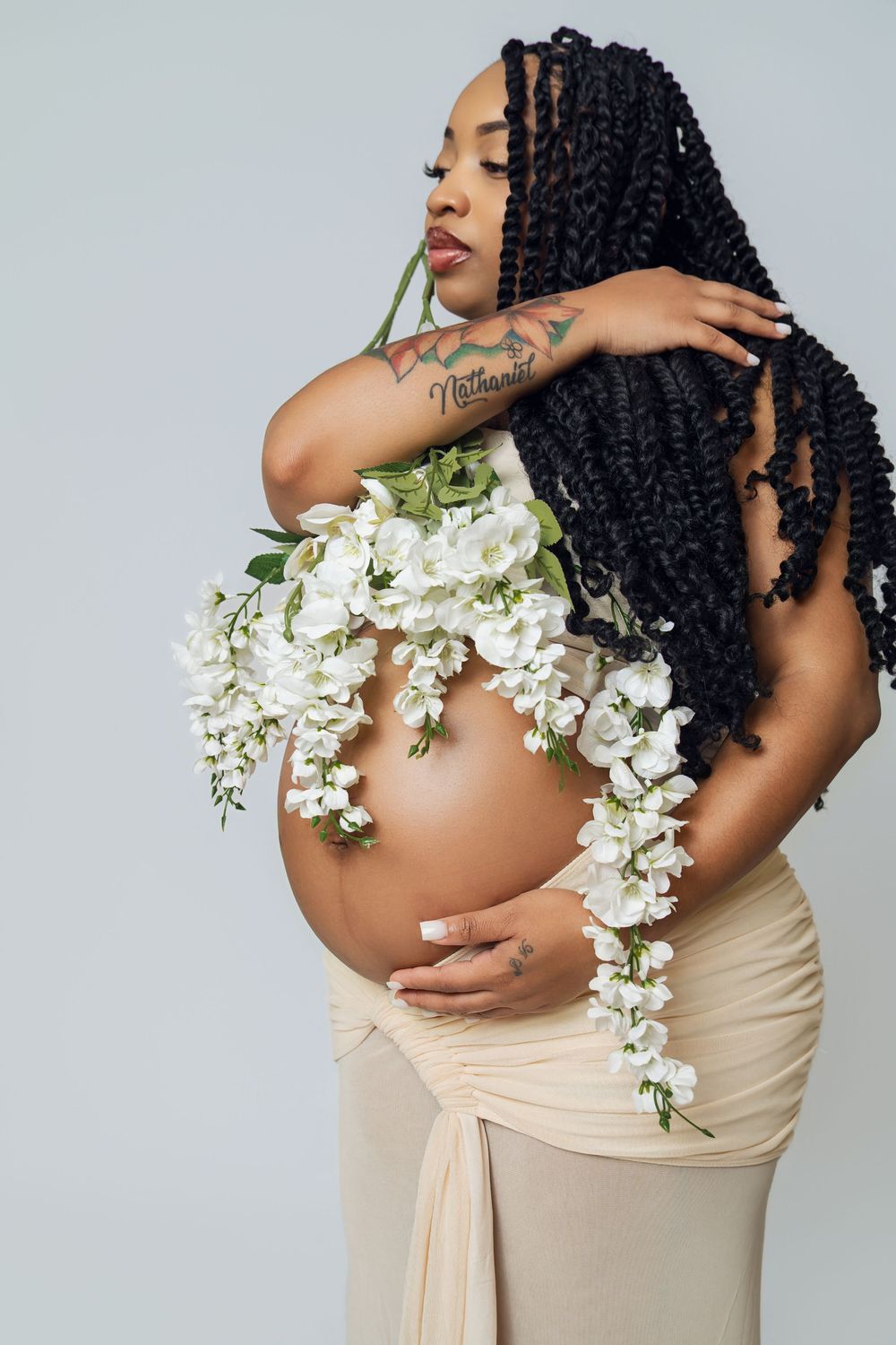 Pregnant Black woman with braids poses with white flowers adorning her bare belly, wearing a beige skirt.