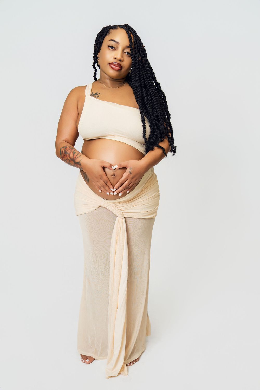 Pregnant Black woman with long braids wearing beige crop top and maxi skirt, cradling baby bump with heart hands.