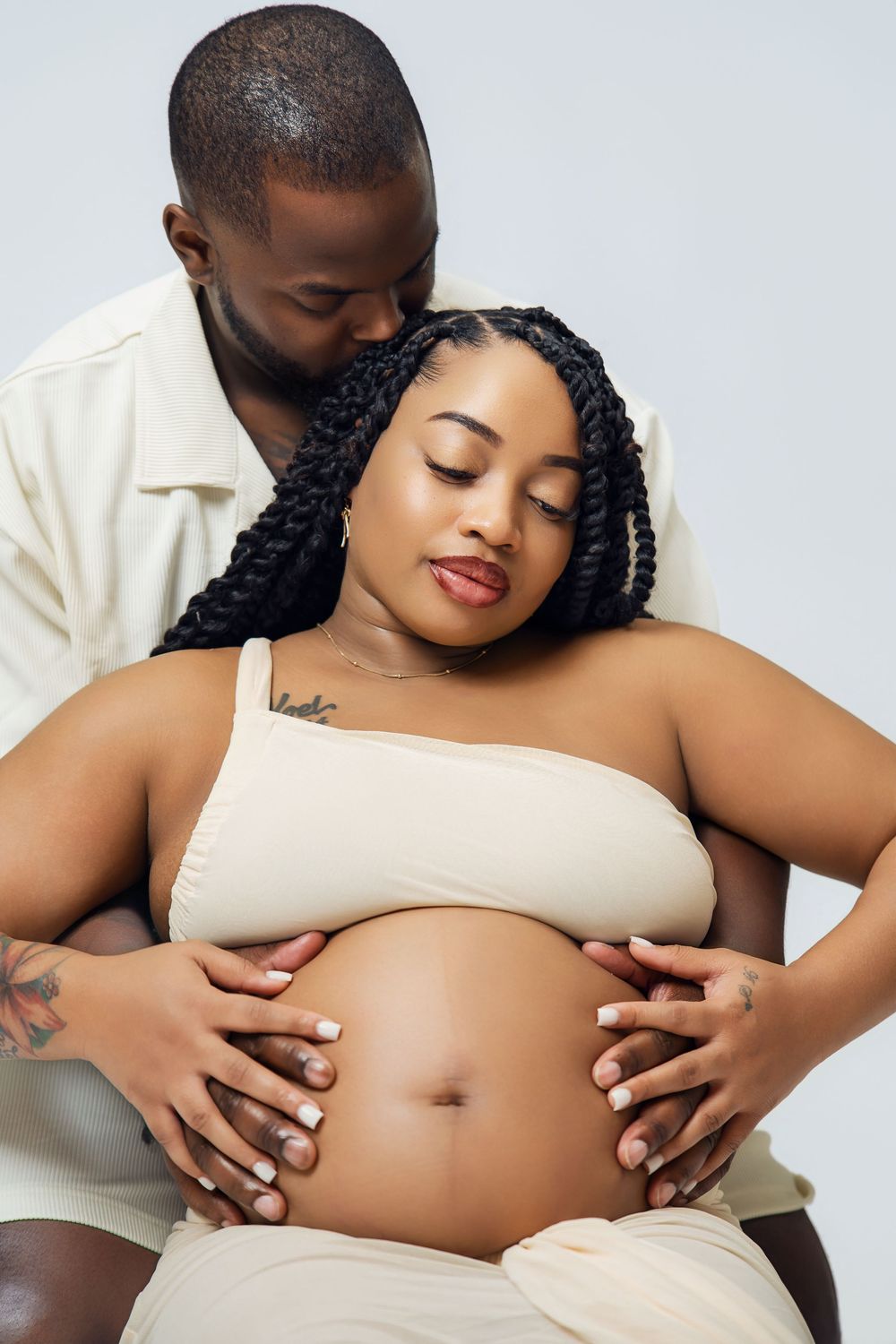 A couple tenderly cradles a pregnant belly, the man kissing her head, both wearing cream outfits against a gray background.