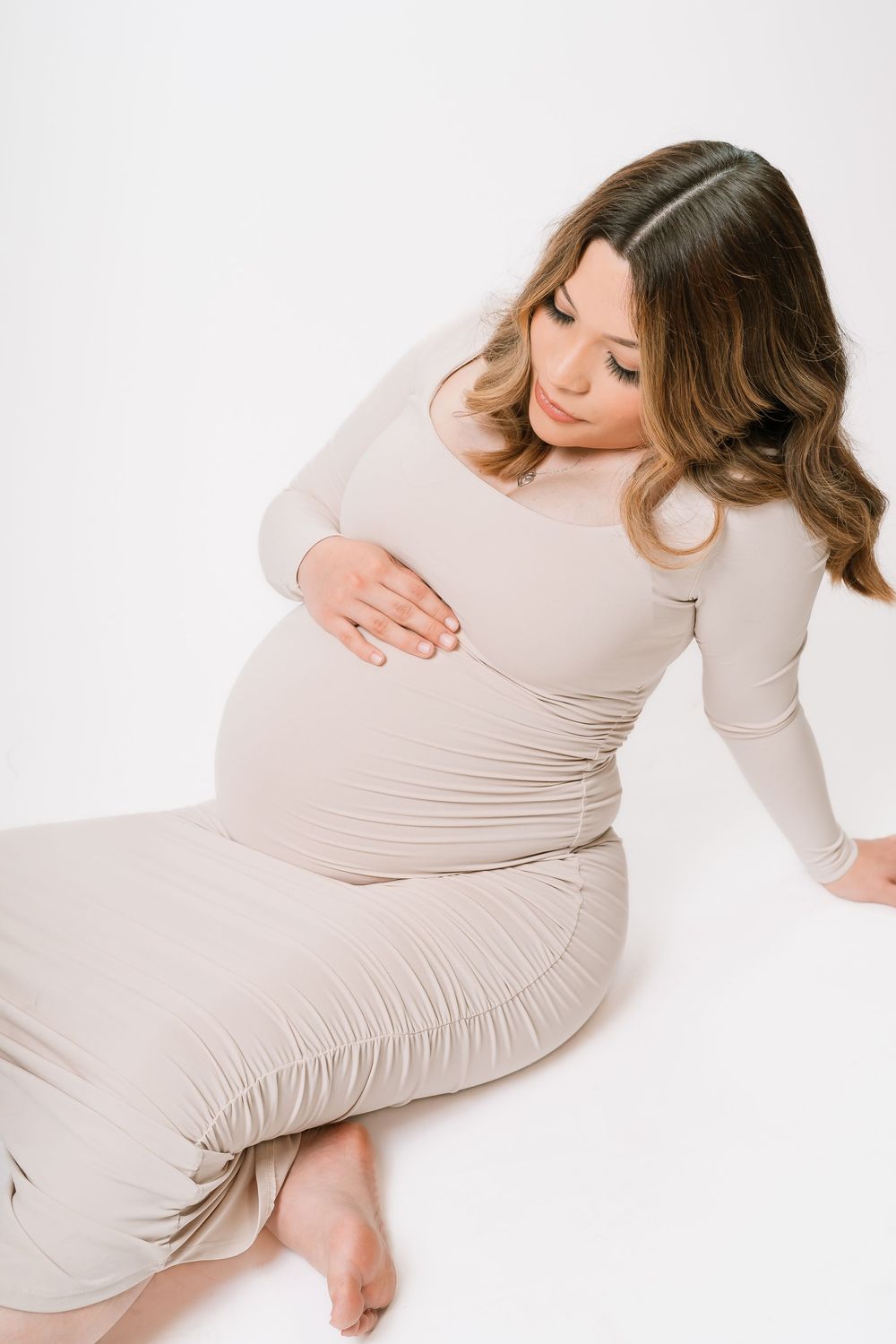 Pregnant woman in beige dress sitting on white background, gently cradling her baby bump with both hands.