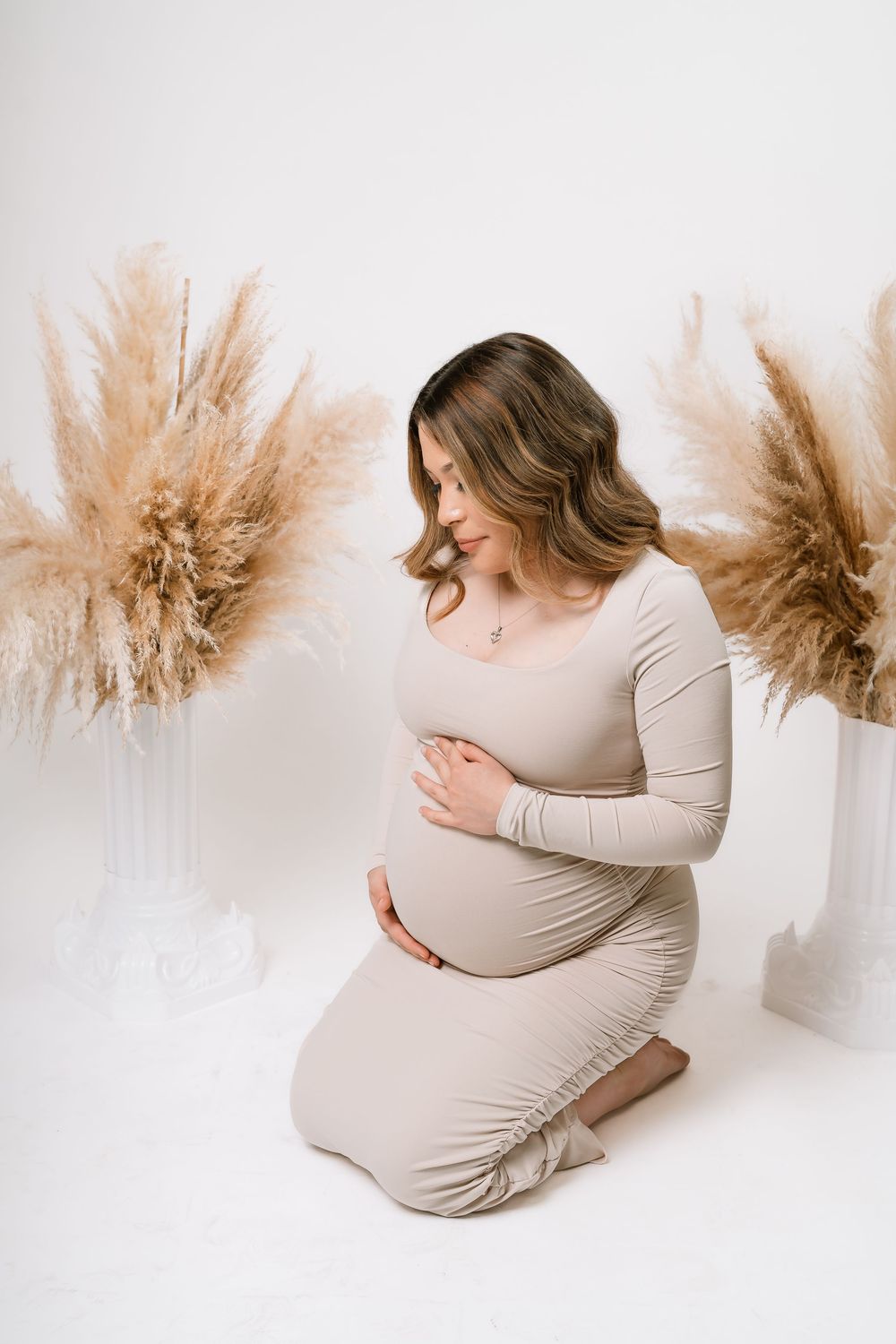 Pregnant woman in beige dress kneeling gracefully, cradling her baby bump, surrounded by pampas grass in white vases.