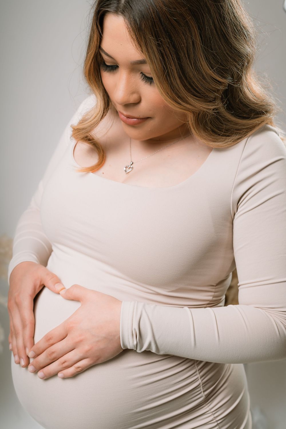 Pregnant woman in beige dress cradling baby bump, wearing a necklace, looking down tenderly.