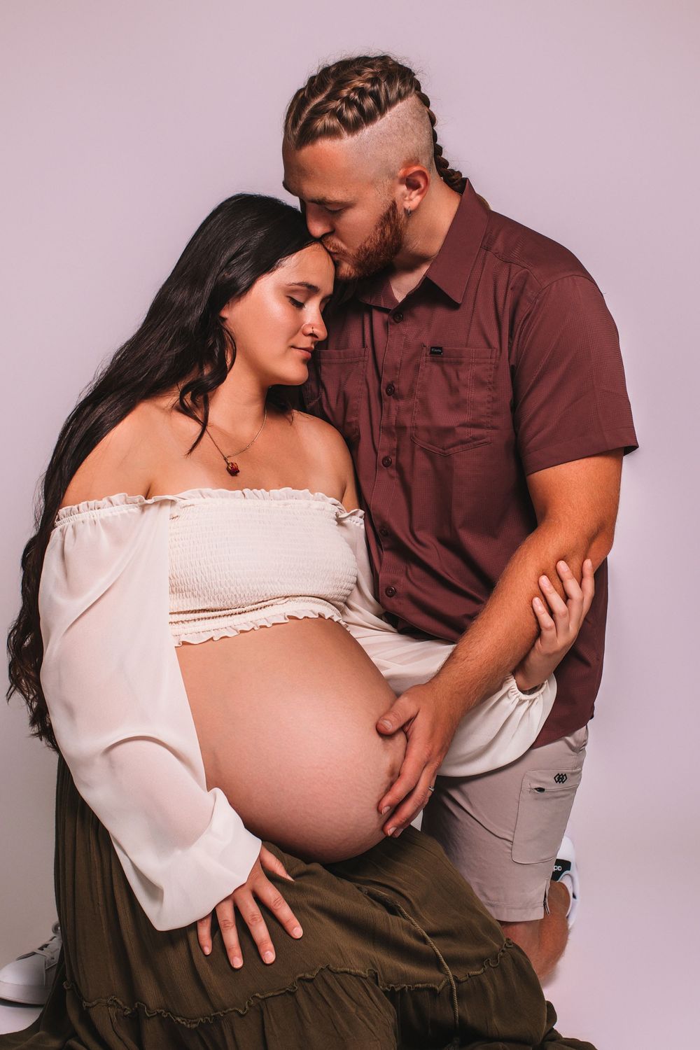 Couple sharing a tender maternity photo shoot moment, man kissing woman's head while cradling her pregnant belly.