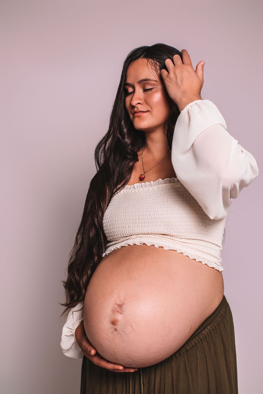 Pregnant woman in white crop top and olive skirt cradling her baby bump with eyes closed against light background.