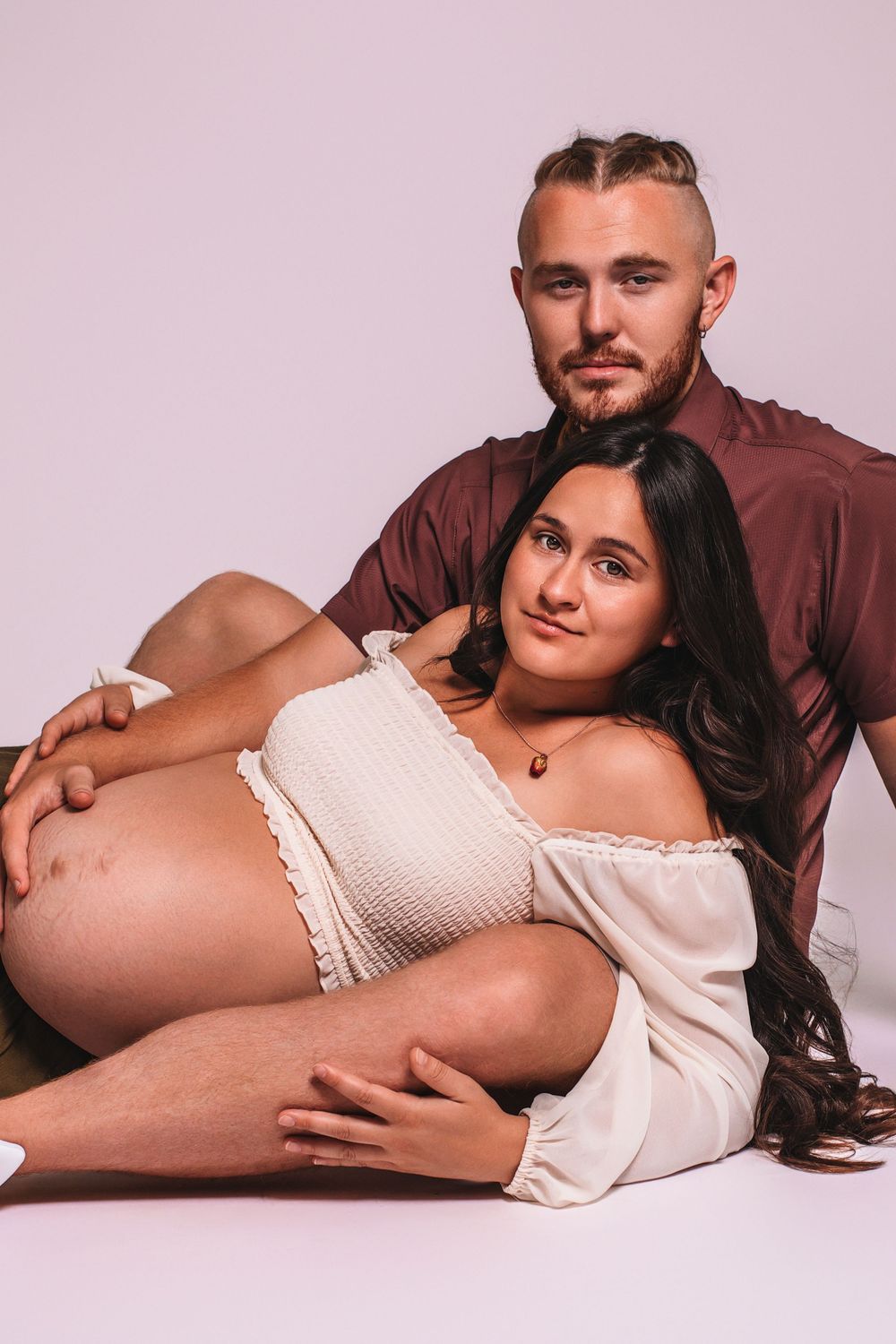 Couple maternity photo with pregnant woman in white off-shoulder dress leaning against partner on white background.