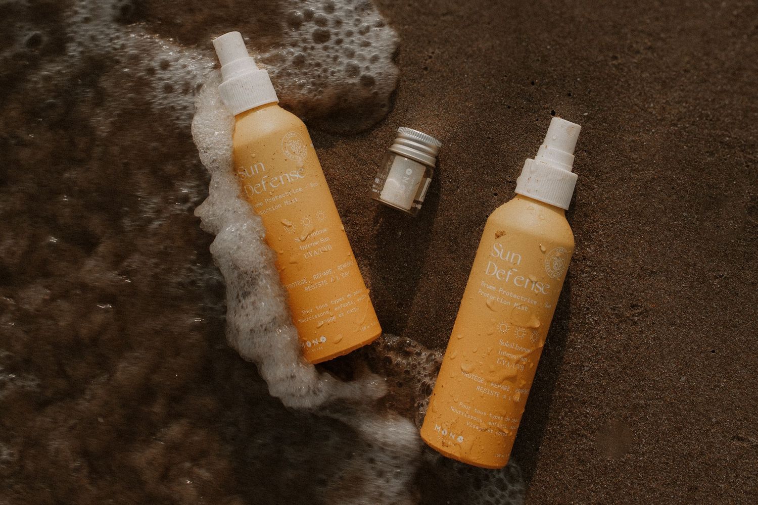 Two orange spray bottles and a small jar of beach skincare products resting on wet sand with ocean foam.