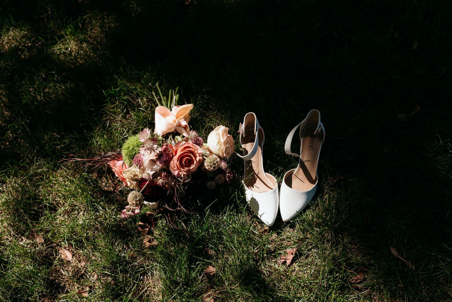Bridal bouquet with roses and white heels on green grass in sunlight.