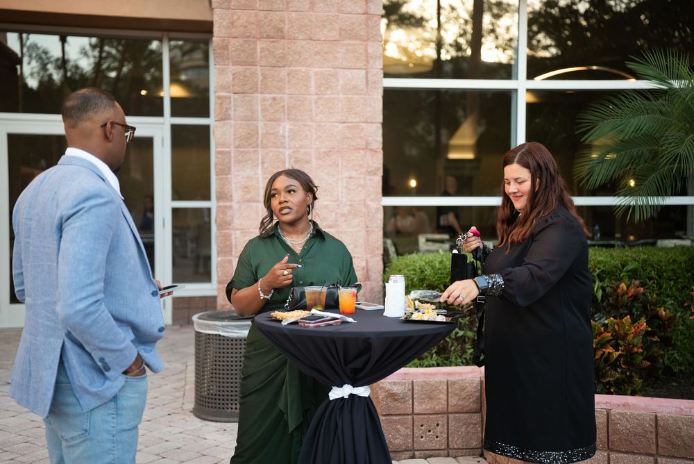 Three professionals networking outdoors at a cocktail reception, standing around a high-top table with food and drinks.