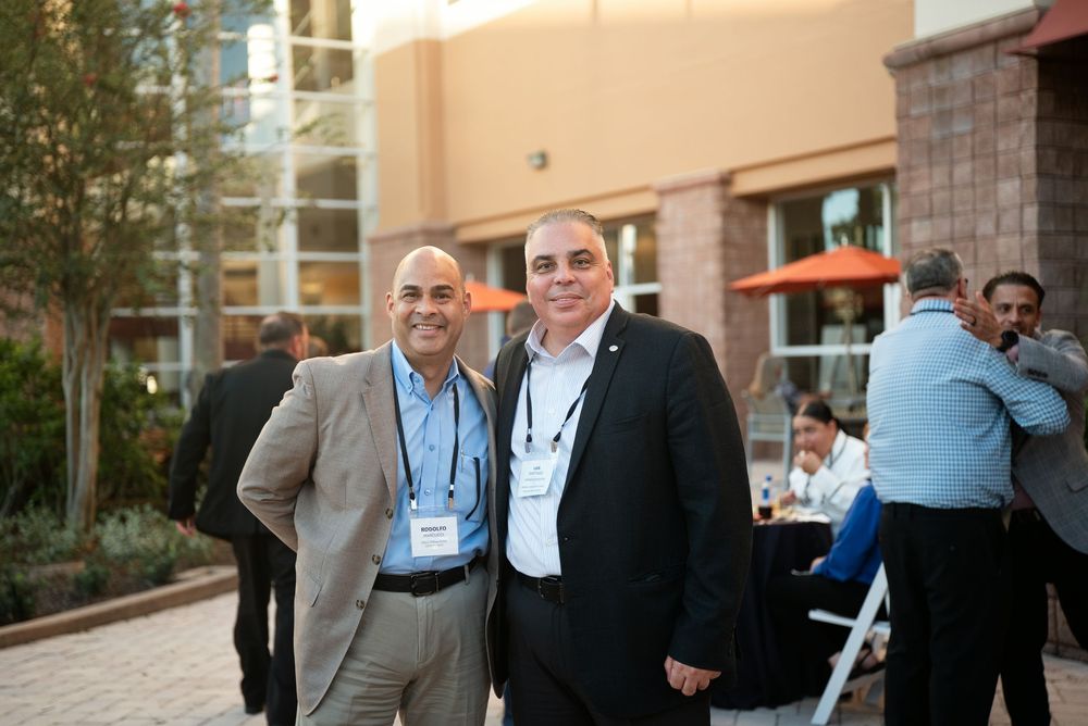 Two men in business attire smile together at an outdoor corporate networking event with guests mingling in the background.