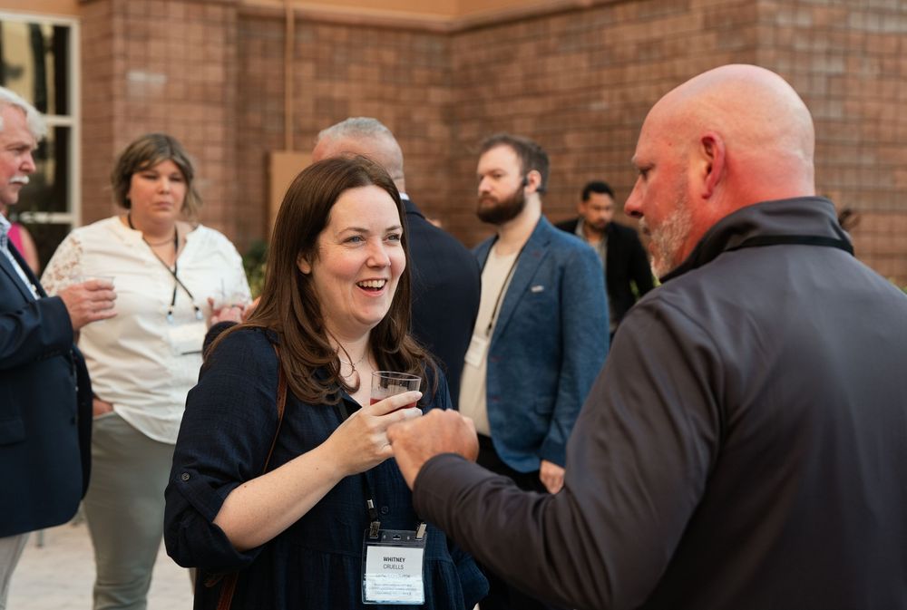 Professionals networking at a corporate event, smiling woman in navy blazer shaking hands with bald man in a brick-walled venue.