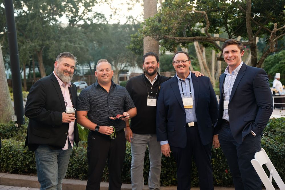 Five businessmen smiling and posing together outdoors at a professional networking event at dusk.