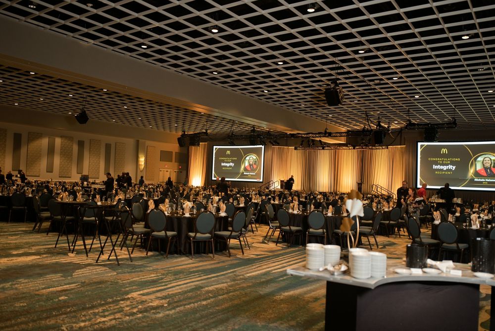 Large corporate gala event in a ballroom with round tables, attendees, and presentation screens displaying logos.