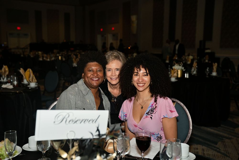 Three women smiling together at a reserved table during a formal banquet event in an elegant ballroom.