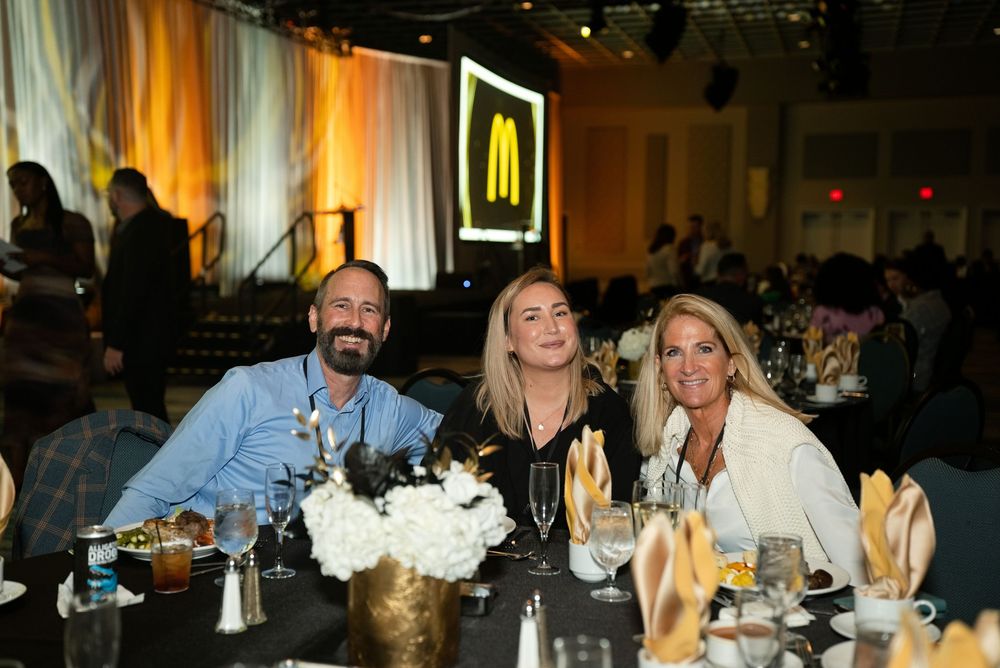 Three guests smile at a gala dinner table with white floral centerpiece, McDonald's logo glowing on stage behind them.