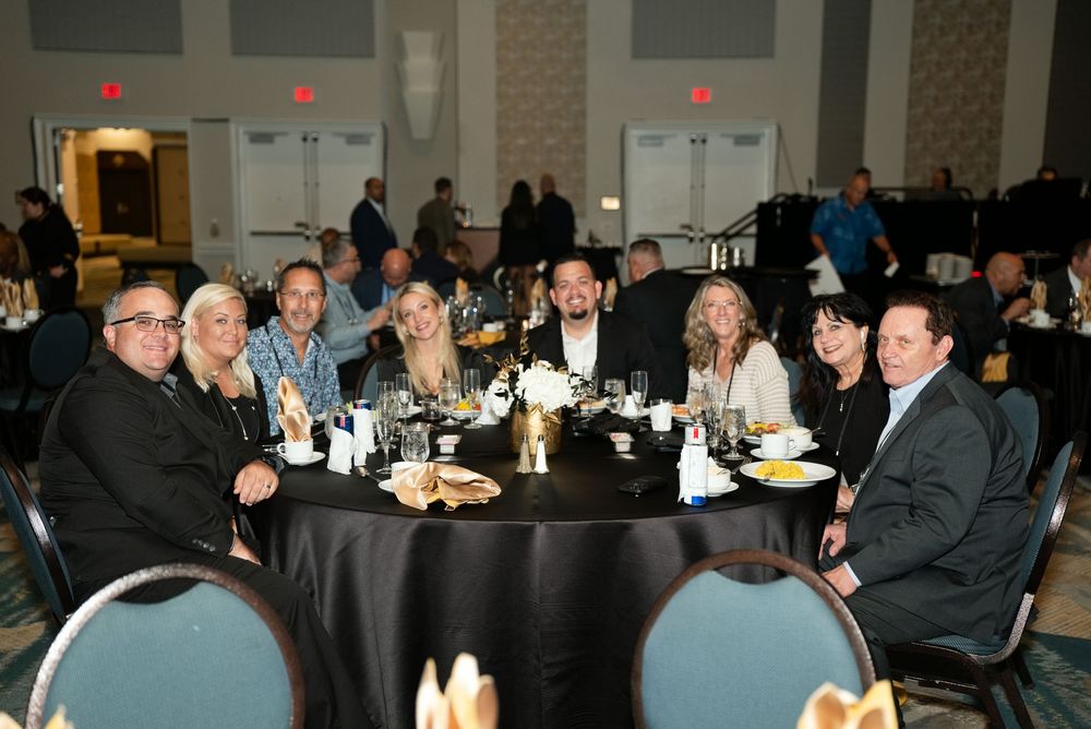 Group of eight adults smiling at a round black-draped table during a formal banquet event in an elegant ballroom.