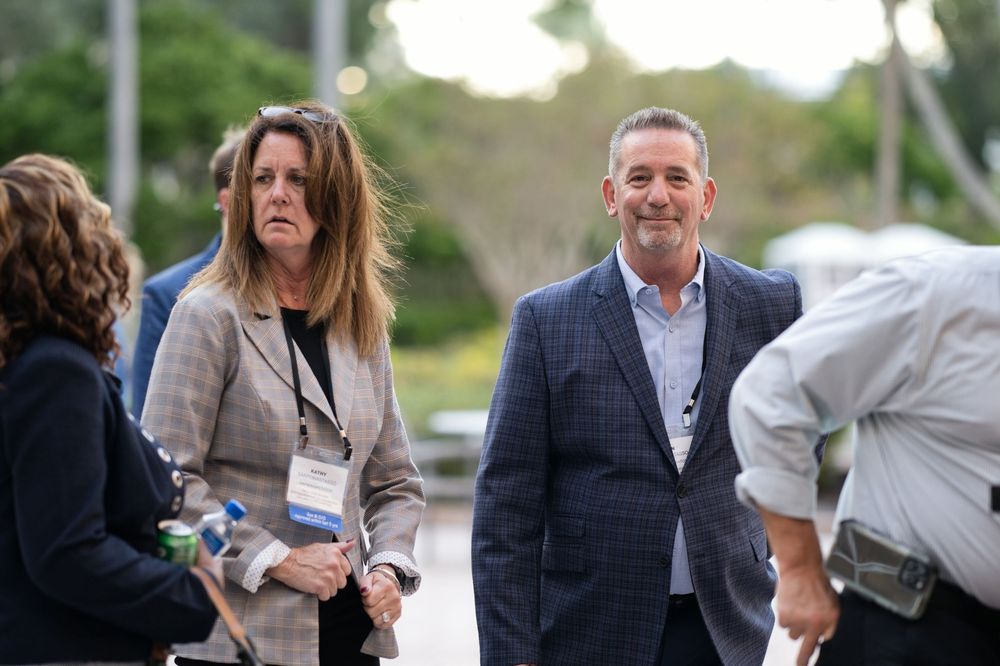 A man in a blue blazer and woman with a lanyard badge walk together at an outdoor professional event.
