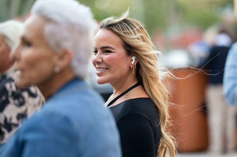 Smiling blonde woman with flowing hair in a black top stands outdoors among a crowd on a sunny day.