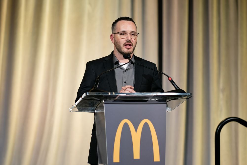 Man in glasses and black blazer speaking at a McDonald's branded podium during a formal event.