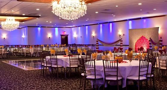 An elegant banquet hall with crystal chandeliers, purple uplighting, and round tables set for an Indian celebration.