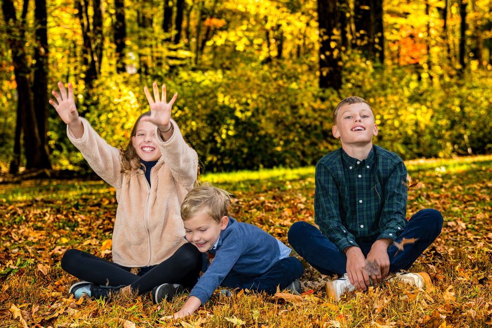 Three happy children sitting on autumn leaves in a park with golden yellow trees in the background.