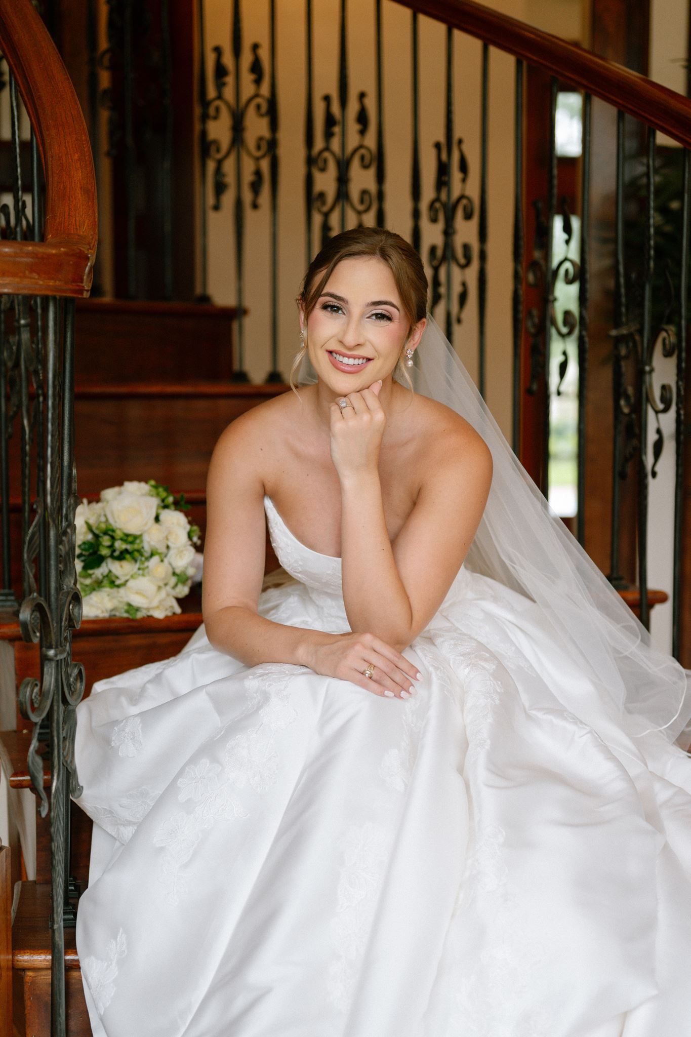 Smiling bride in strapless white gown and veil sits on wooden staircase holding white bouquet.