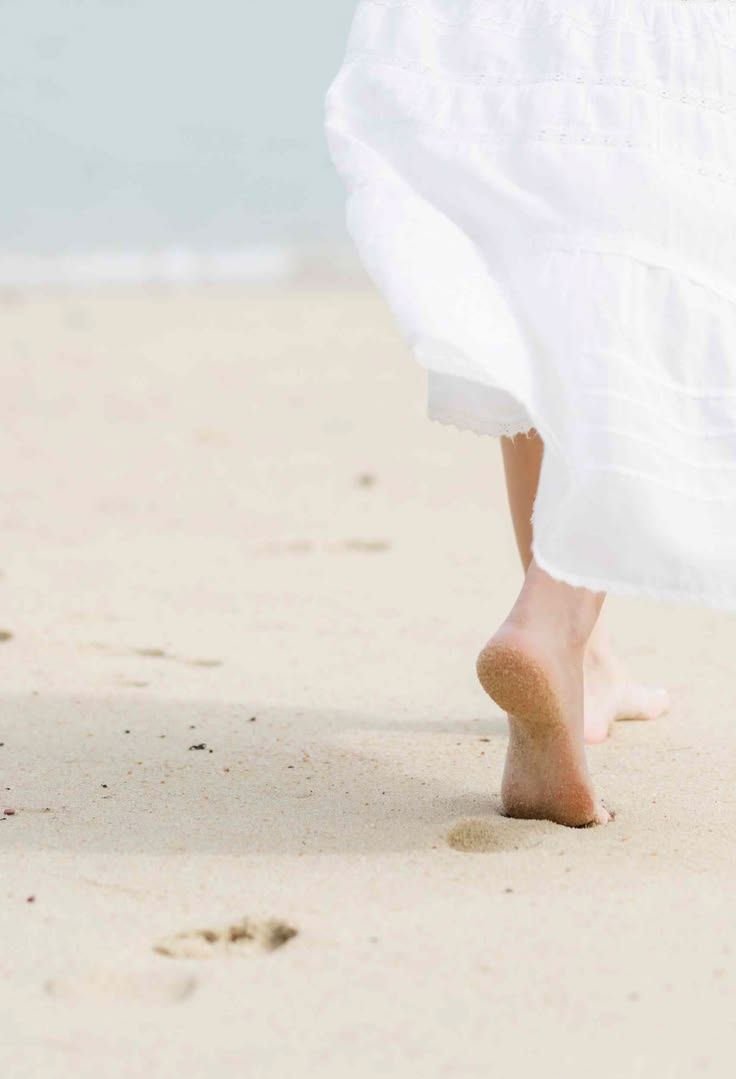 Barefoot woman in white dress walking on sandy beach, leaving footprints in the sand near ocean shore.