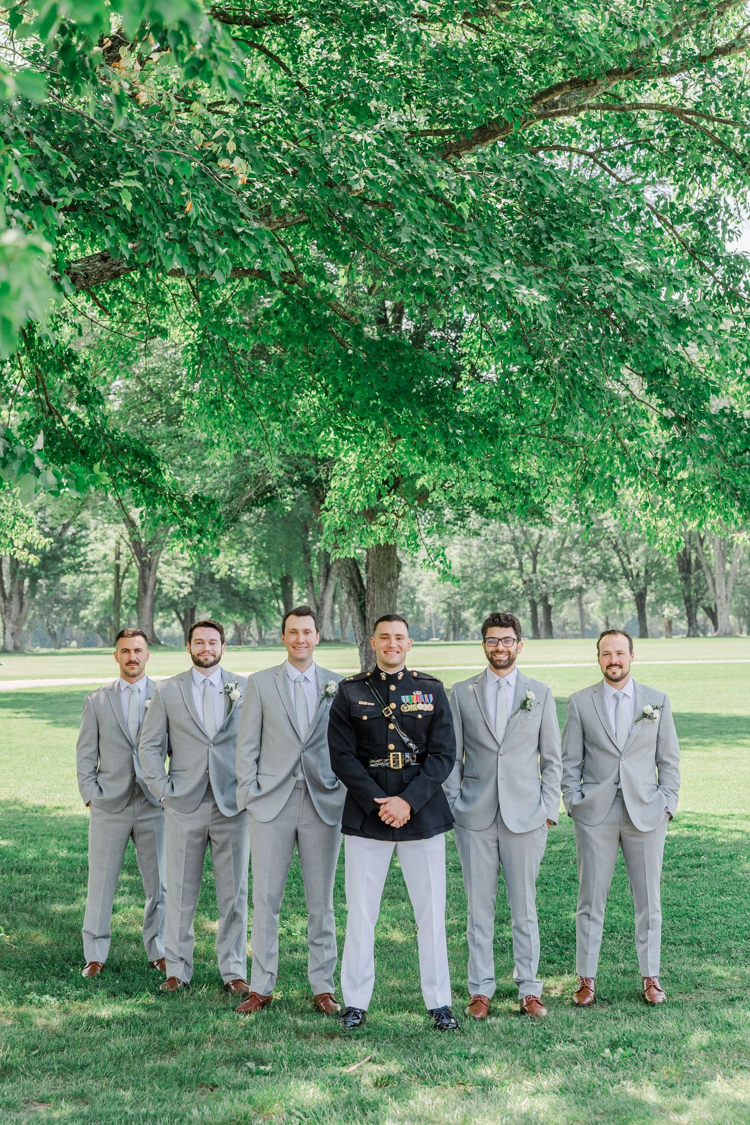 Marine groom in dress uniform stands with groomsmen in gray suits outdoors under lush green trees.