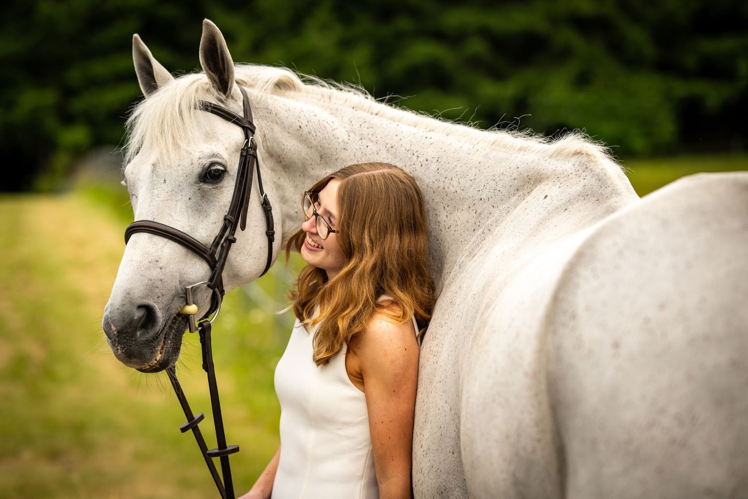 A smiling young woman in a white dress stands closely beside a white horse with a bridle in a green outdoor setting.