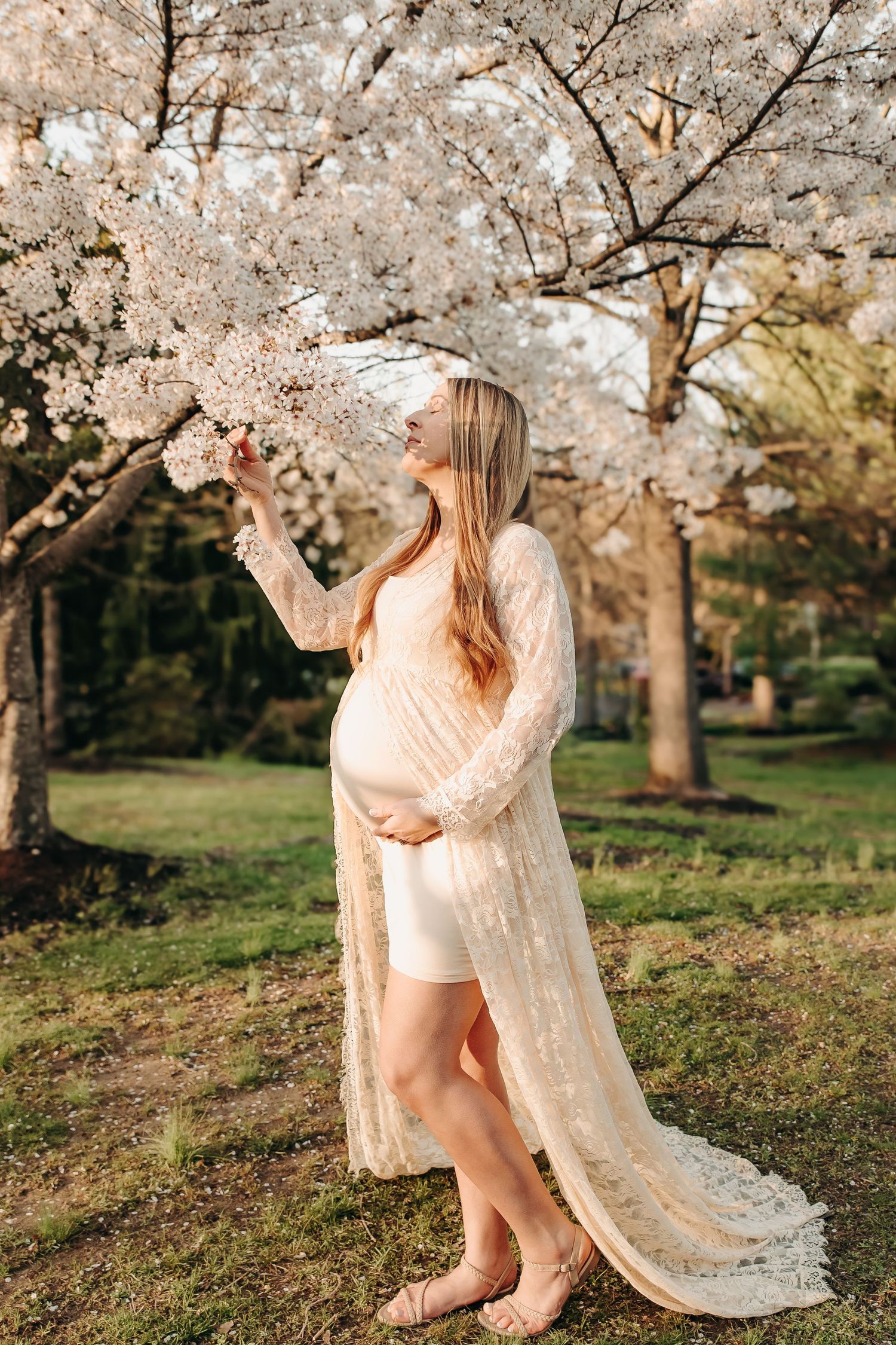 Pregnant woman in cream lace duster touching cherry blossoms in a sunlit park, cradling her baby bump.
