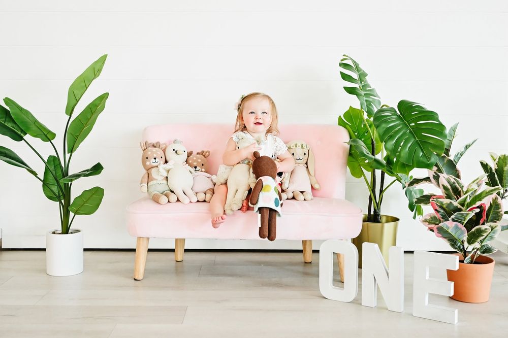 Adorable baby sitting on a pink couch with stuffed animals beside an ONE sign and green plants.