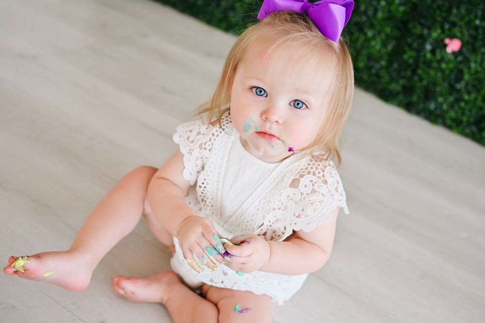 Cute blonde baby girl with blue eyes wearing a white lace outfit and purple bow headband.