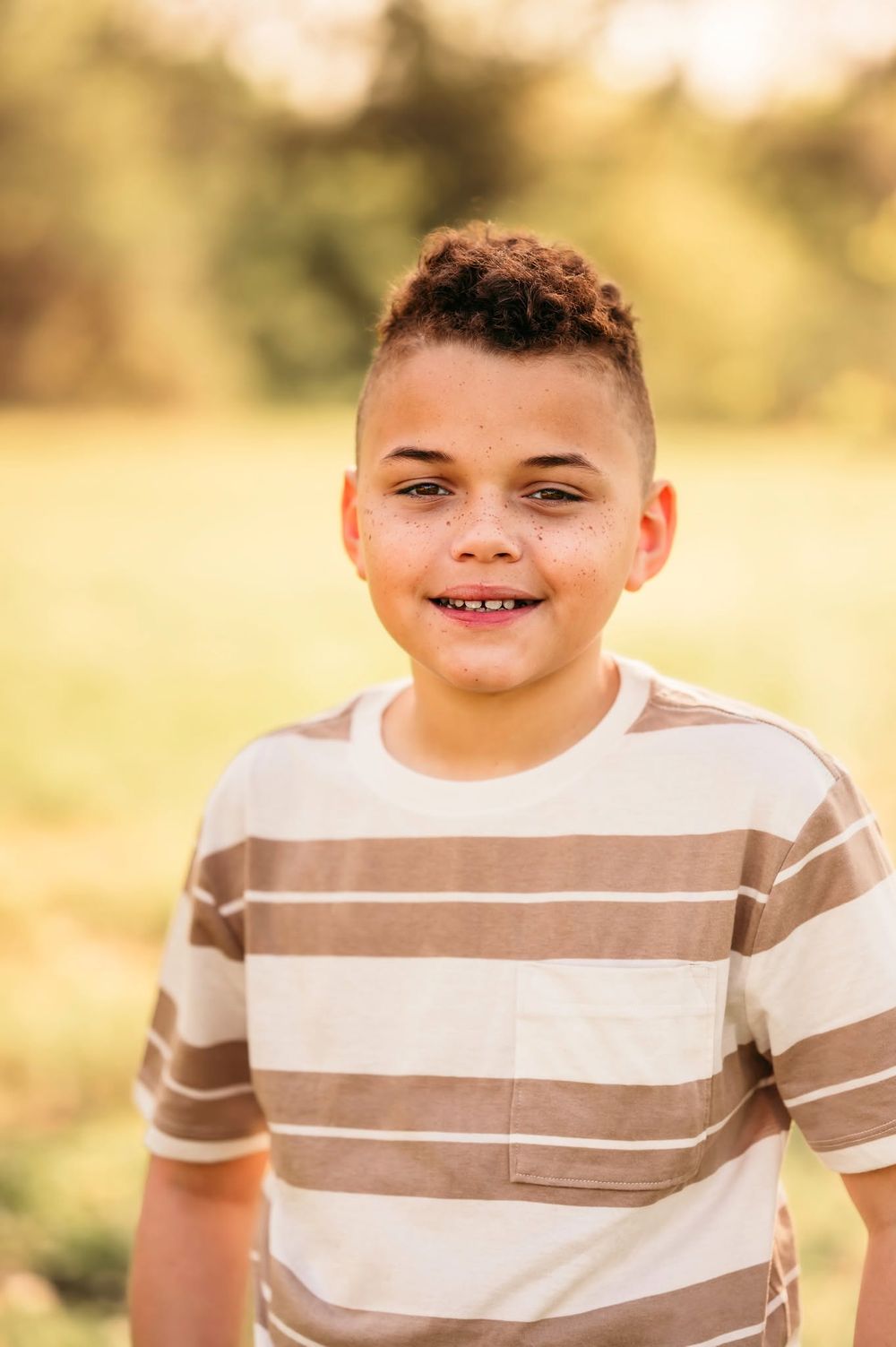 Smiling young boy with freckles wearing a striped t-shirt in a sunny outdoor setting.