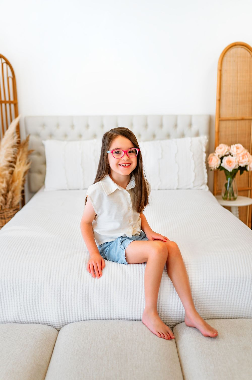 Cheerful girl in pink glasses and denim shorts sitting on edge of white bed.