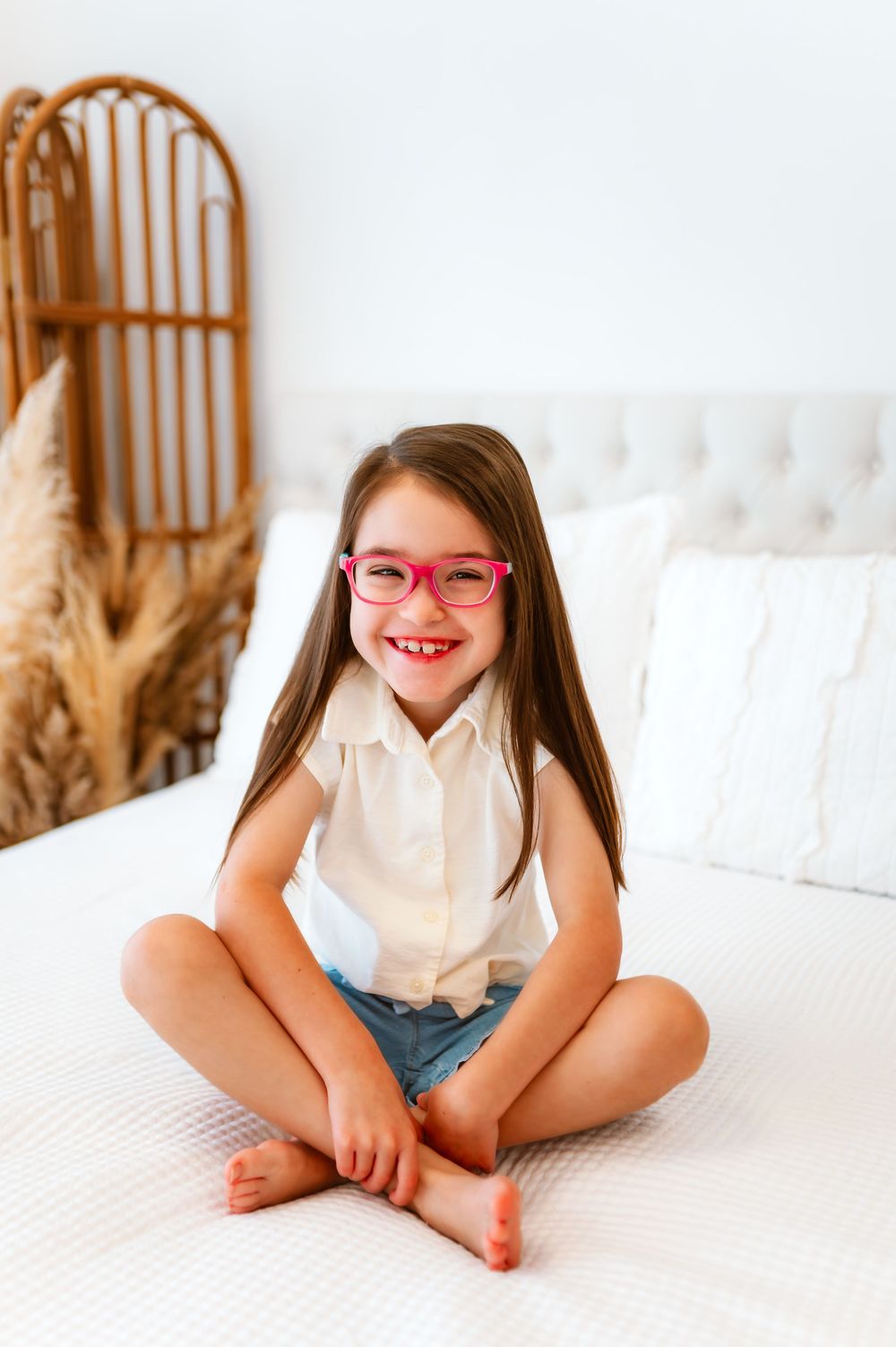 Happy young girl with pink glasses sitting cross-legged on white bed, smiling brightly.