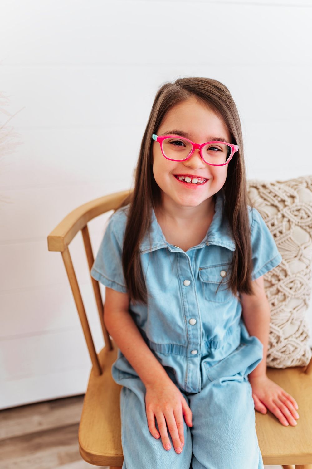 Smiling girl with pink glasses in denim jumpsuit sitting on wooden chair indoors.
