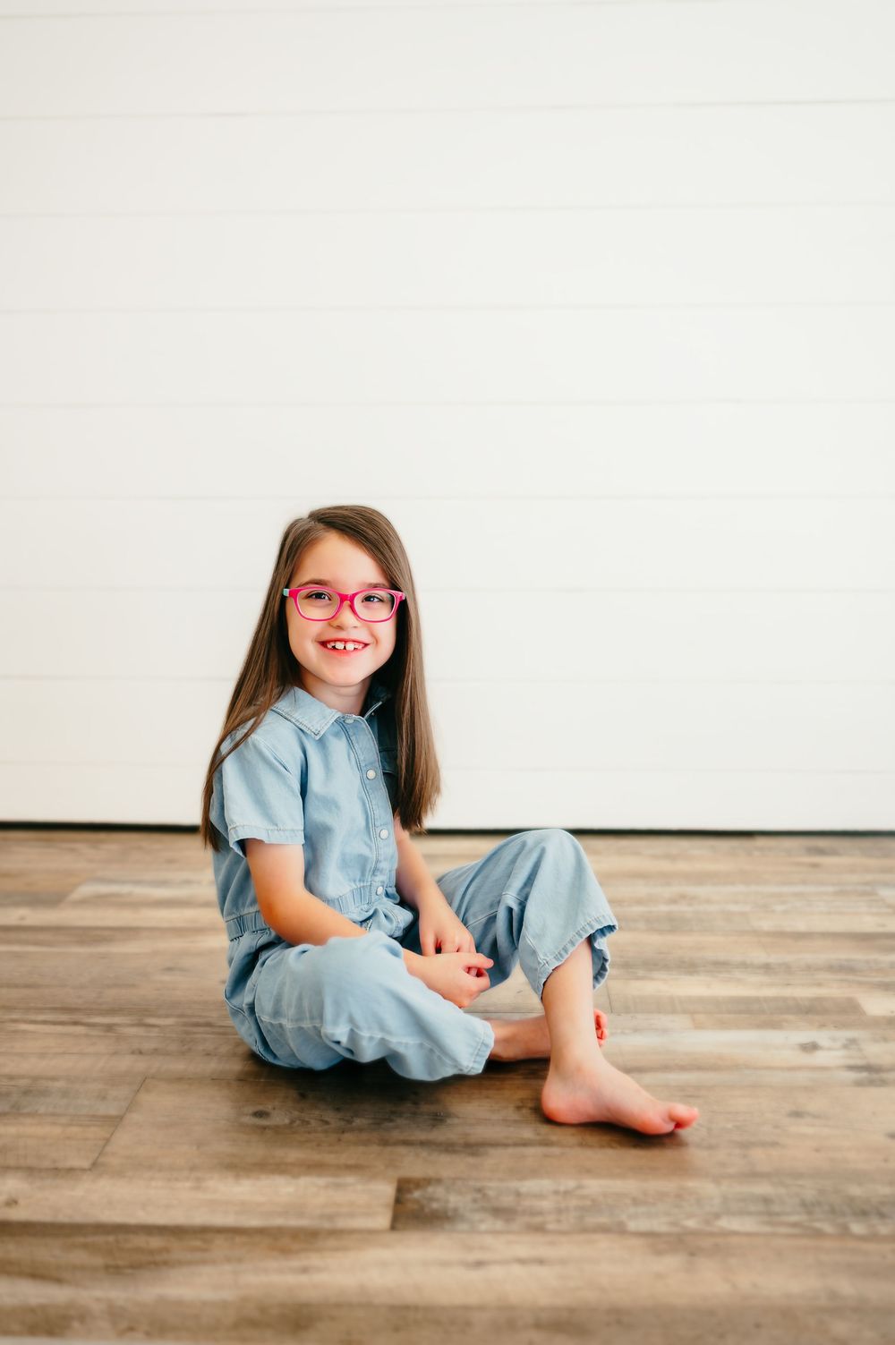 Happy girl in pink glasses and denim jumpsuit sitting on hardwood floor, smiling.