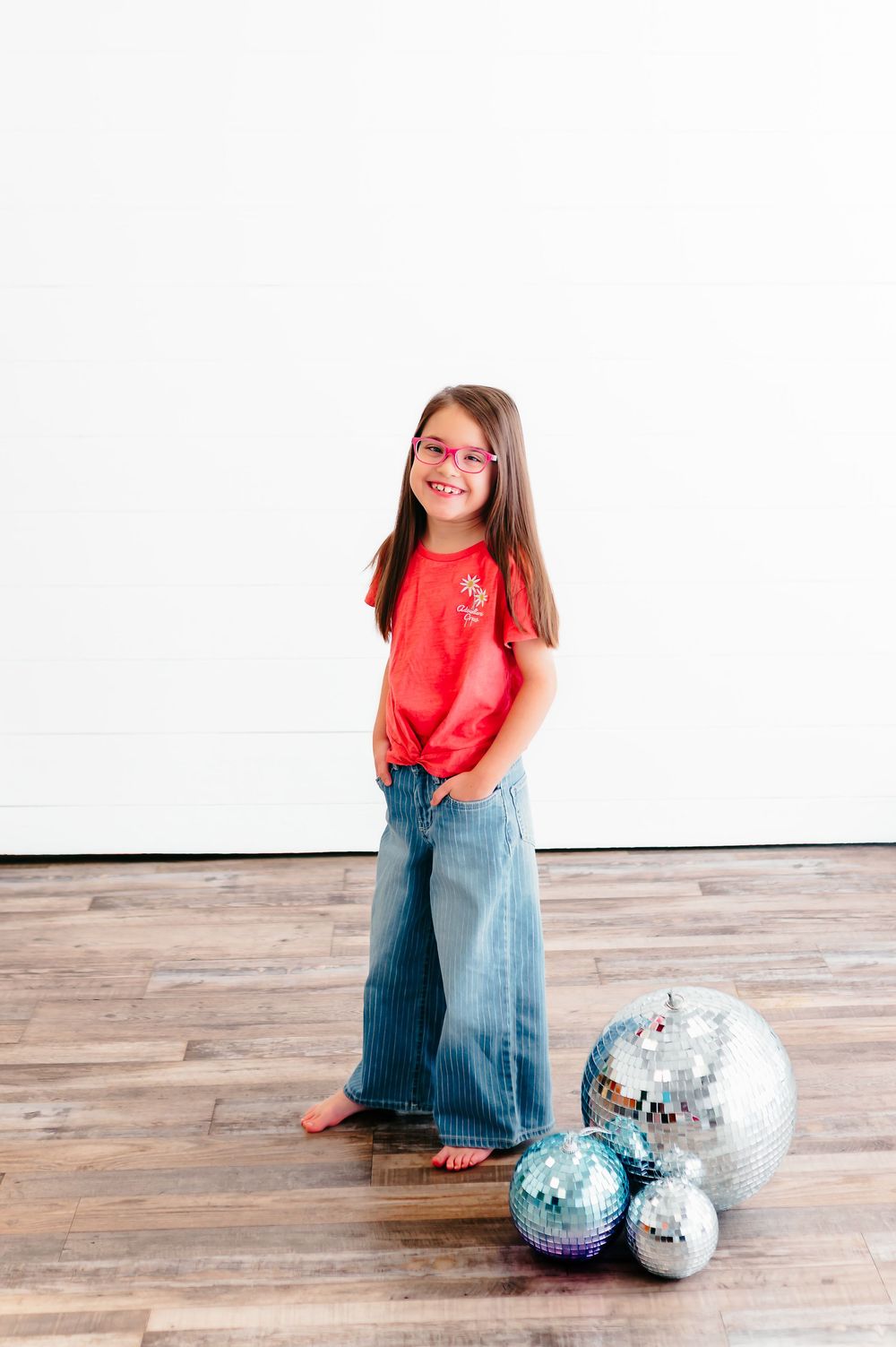 Young girl in red shirt and wide-leg jeans posing with silver disco balls indoors.