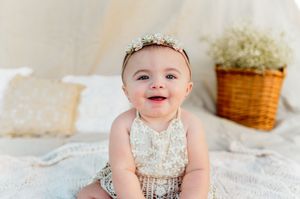 Adorable smiling baby in lace romper with floral headband indoors.