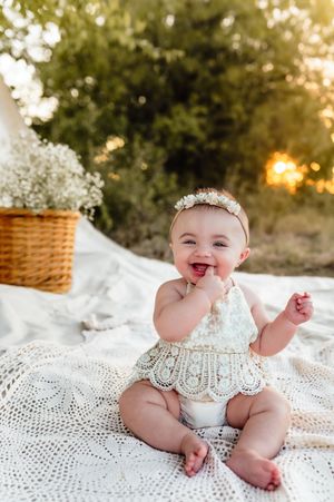 Smiling baby in lace romper sitting on blanket outdoors at sunset.