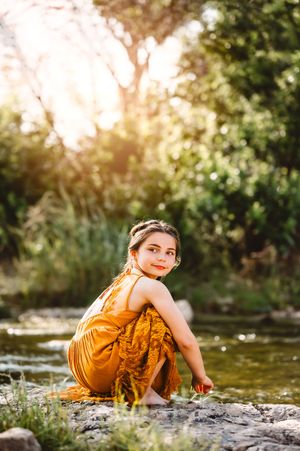 Young girl in a golden yellow dress sitting by a creek surrounded by lush green trees in sunlight.